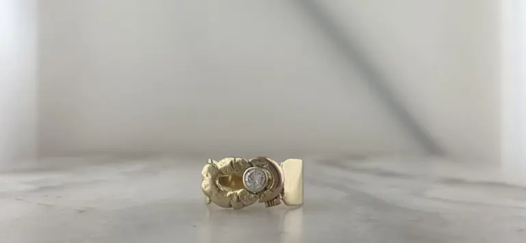 Close-up of a gold ring with a clustered design and a central diamond, resting on a marble surface against a plain white background.