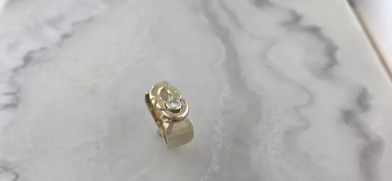 A gold ring with a central gemstone, possibly a diamond, and decorative elements, placed on a white textured surface
