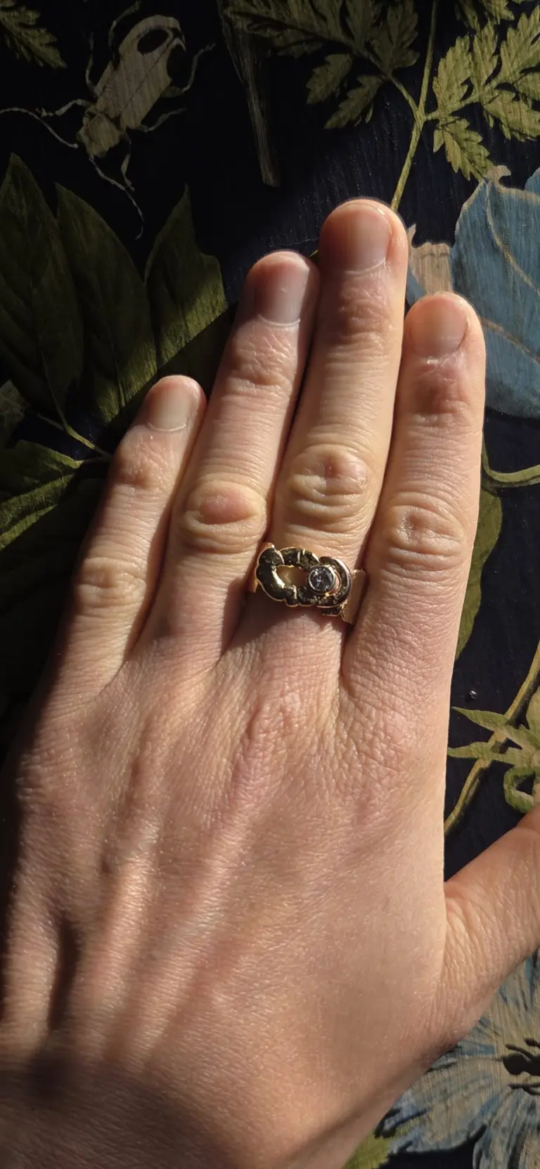 A close-up of a person's left hand wearing a ring with a gemstone, resting on a patterned fabric background.