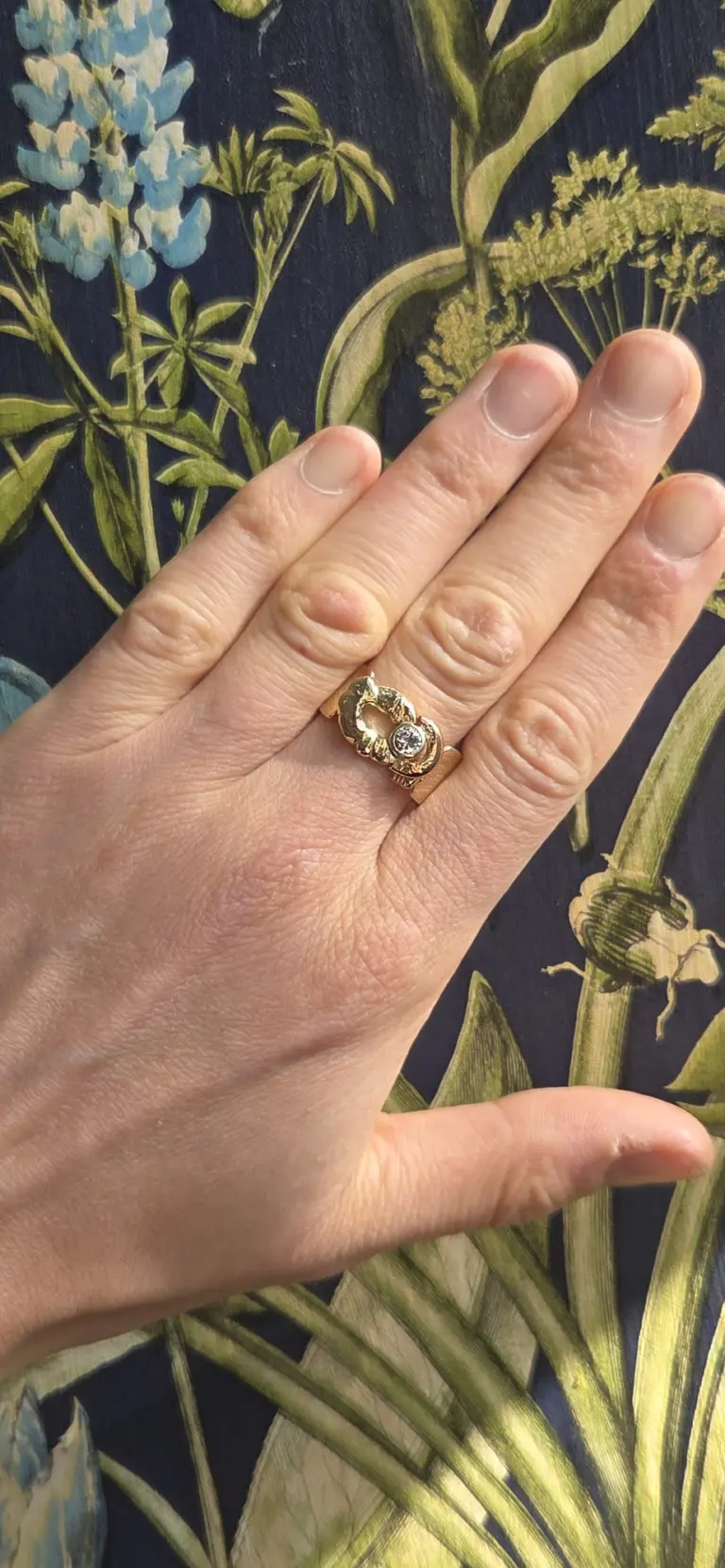 Close-up of a person's hand with a gold ring on the ring finger, featuring a central gemstone and a moon-shaped design, against a blurred green foliage background