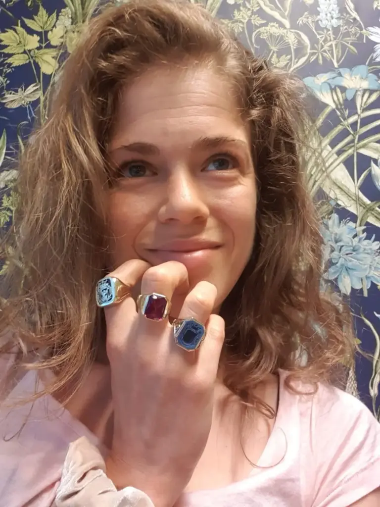 A woman with curly brown hair posing in front of a wallpaper with botanical designs, wearing rings on her fingers and a light pink top.