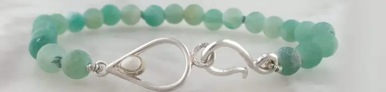Close-up photo of a beaded bracelet with a silver clasp, featuring round pale green and blue beads with a textured surface, on a light surface background.
