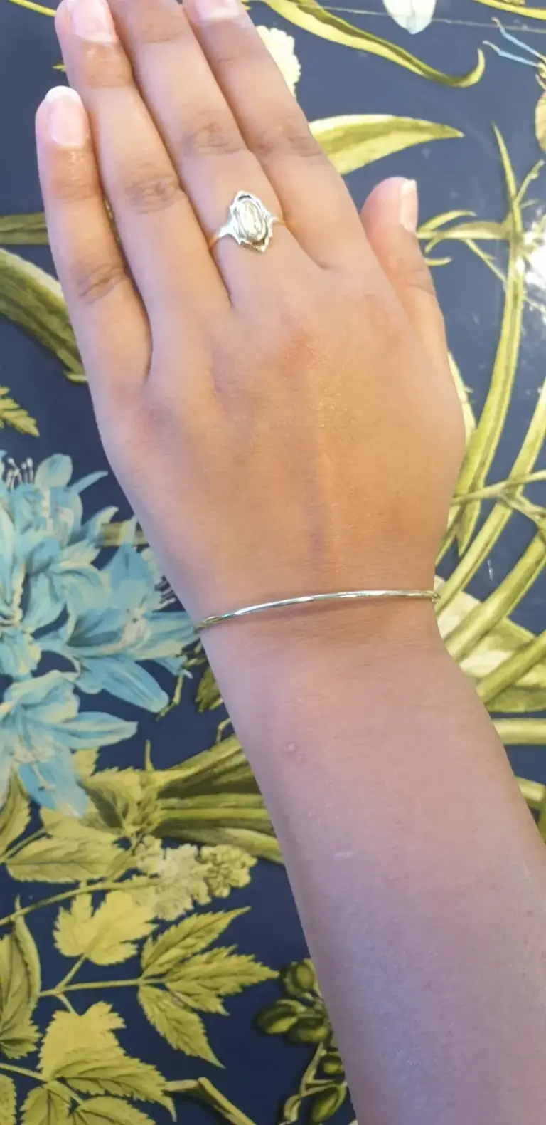 A hand with a silver ring on the middle finger and a silver bracelet on the wrist rests on a floral patterned surface with blue, green, and yellow flowers and leaves.