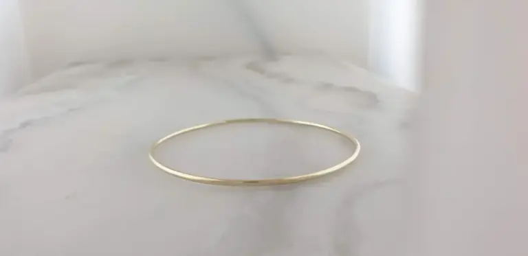 A simple gold ring bracelet resting on a white marble surface with gray veins, in front of a plain gray background.