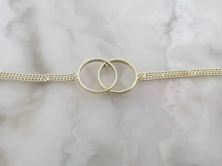 Close-up of two gold wedding rings intertwined on a silver chain, resting on a white textured surface.