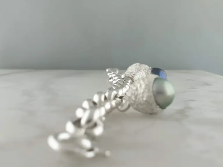 Close-up of jewelry pieces, including a pearl necklace, a textured silver bracelet, and an oval-shaped bead, arranged on a light-colored surface with a plain background.