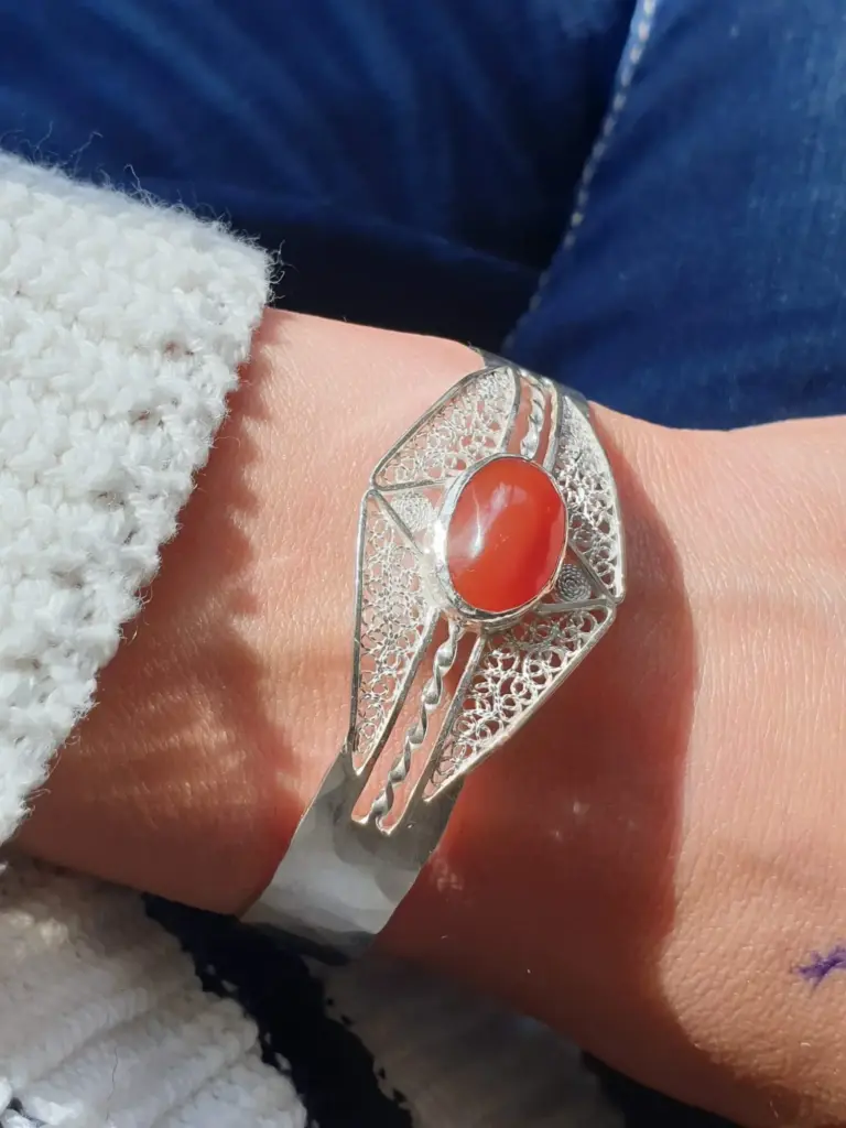 A person wearing a silver ring with a large red-orange stone, showing their wrist and part of their arm. The person has light skin and is wearing a fluffy white or cream-colored sweater, with denim jeans visible in the background.