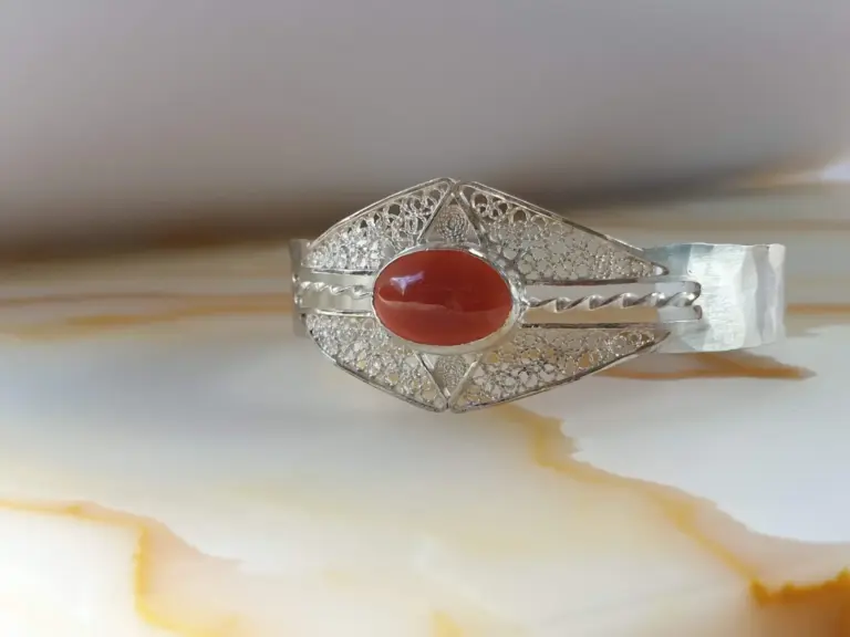 A silver ring with intricate filigree design and a large oval red gemstone in the center, placed on a white surface with subtle yellowish veins.