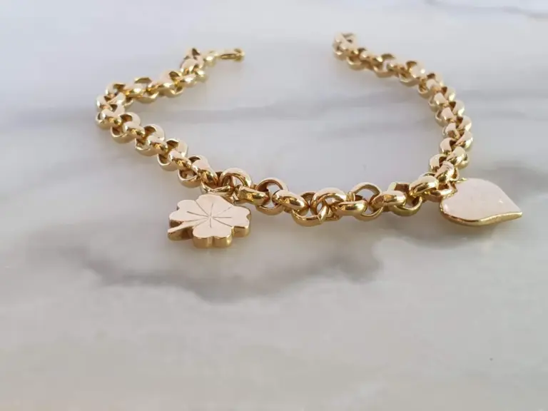 Gold chain bracelet with two charms, one shaped like a four-leaf clover and the other like a flower, lying on a white marble surface.