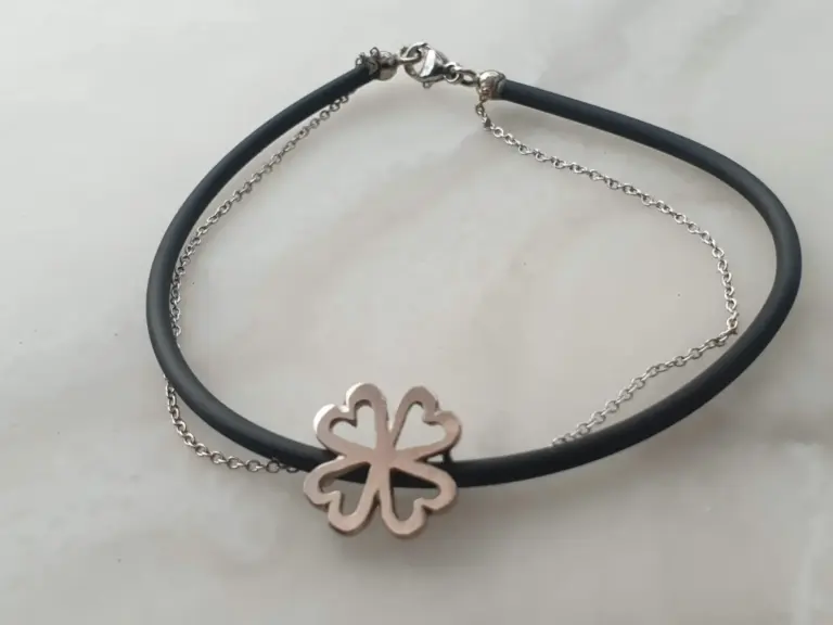A close-up of jewelry necklaces, including a black cord with a silver clasp, a silver chain, and a pinkish flower-shaped pendant, on a light background.