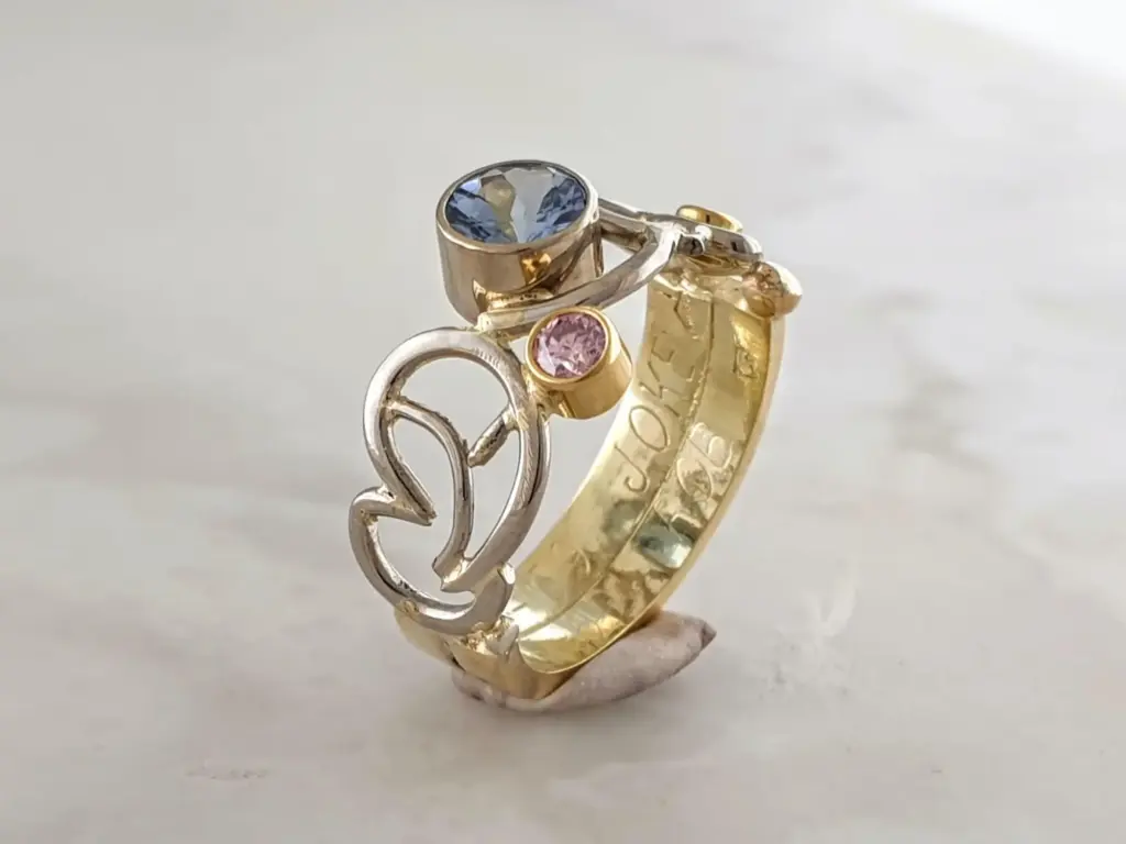 Close-up of a gold ring with multiple intertwined bands, featuring blue and pink gemstones, set against a plain light background.