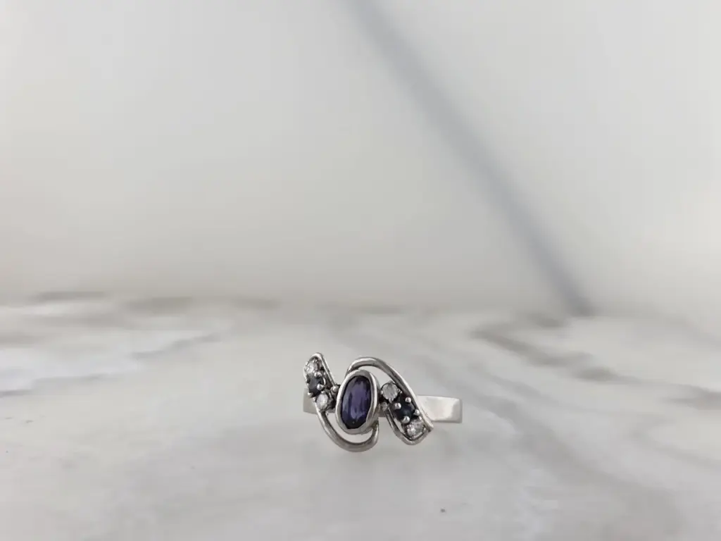 Silver ring with a large oval purple gemstone and smaller clear gemstones, on a white surface with jewelry in the background.