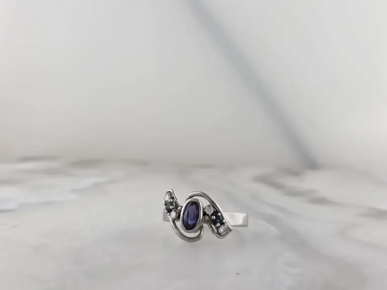 Silver ring with a large oval purple gemstone and smaller clear gemstones, on a white surface with jewelry in the background.