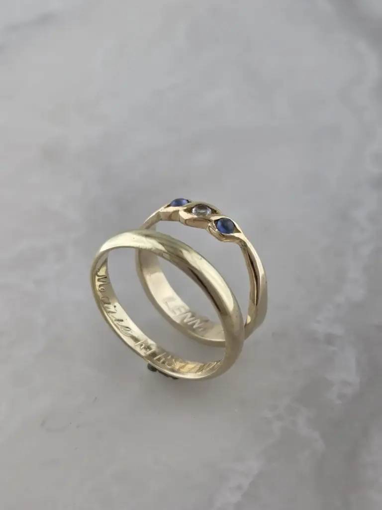 Close-up of two gold rings with engraved text, one with embedded blue and clear gemstones, on a gray surface.
