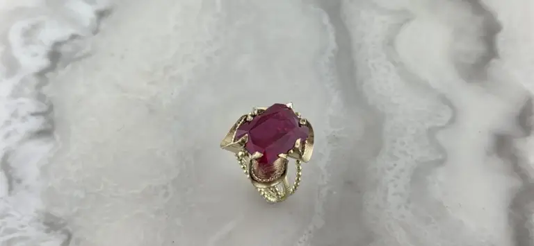 Silver ring with a large pink gemstone in the center, surrounded by intricate detailing and small chains, placed on a white surface.