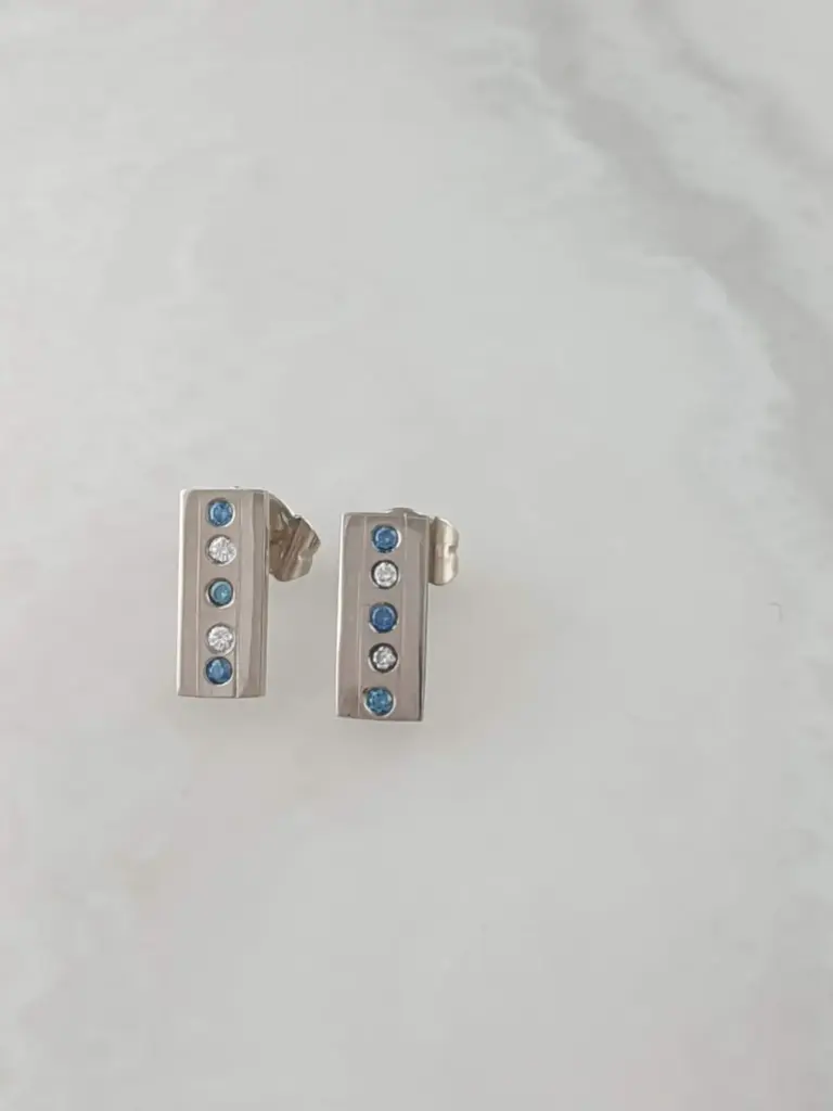 Two small metallic objects with blue and clear gemstones, possibly jewelry pieces, placed on a white surface.