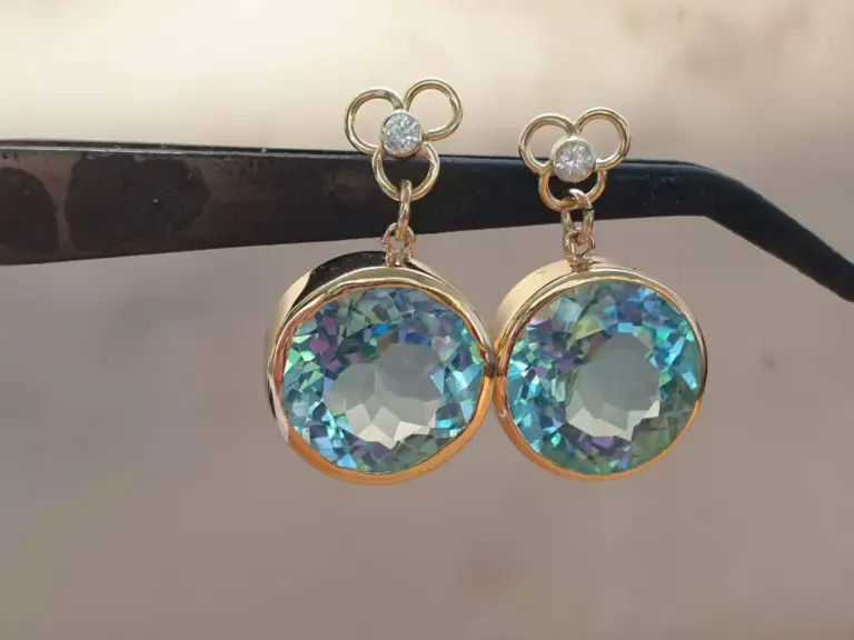 Pair of gold earrings with large circular rainbow crystal pendants and small flower-shaped studs with clear gemstones, hanging from a black textured horizontal bar.