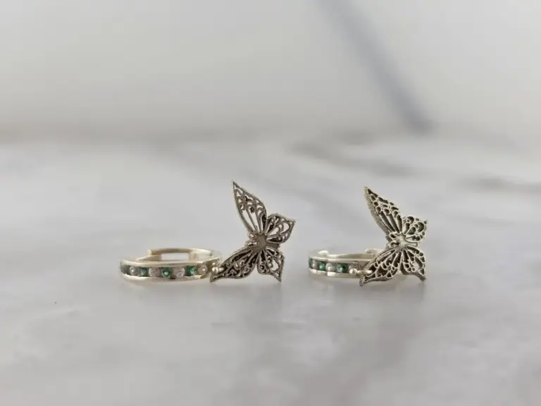 Two rings each featuring a butterfly-shaped decoration, with one ring having a band of alternating green and pink stones and the other with a band of small gemstones.