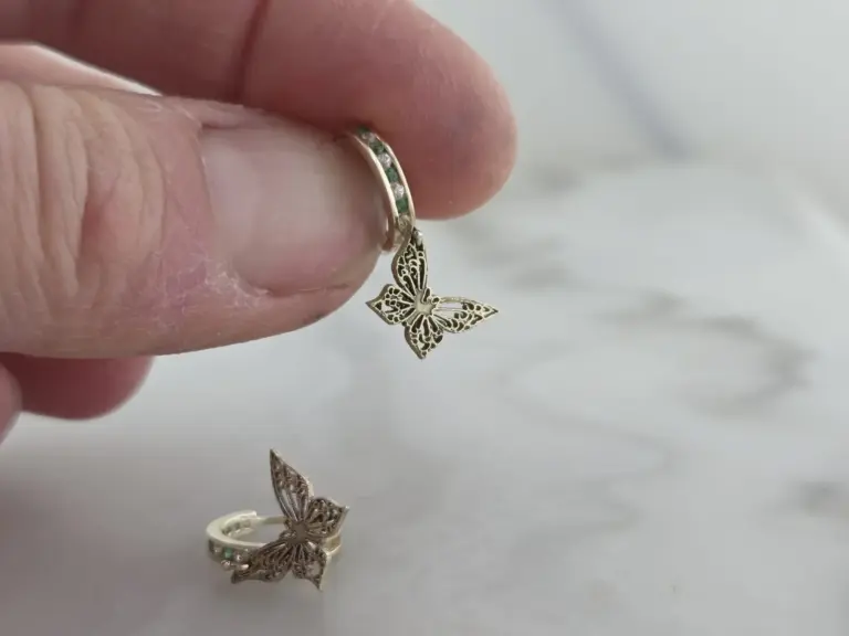 Close-up of a hand holding a butterfly-shaped gold ring with emerald and diamond accents, with other butterfly jewelry on a white surface.