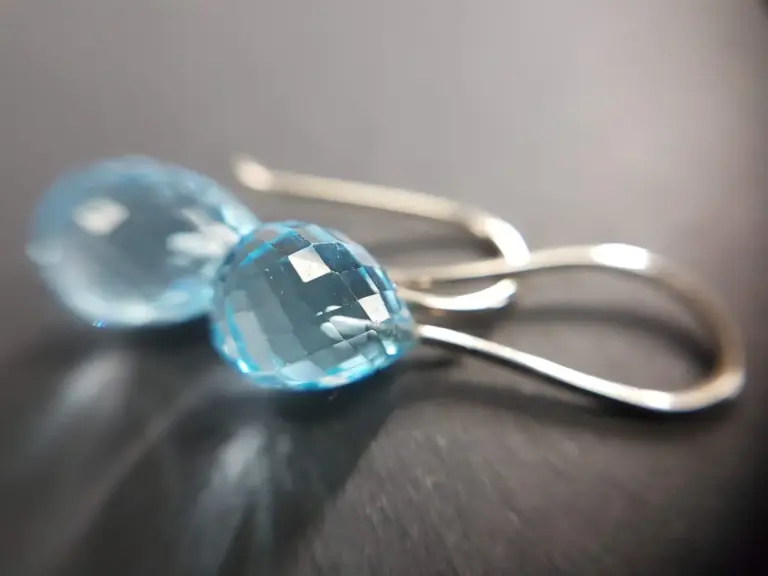 Close-up of a small, faceted, light blue gemstone or glass bead resting on a dark surface with silver tweezers in the background.
