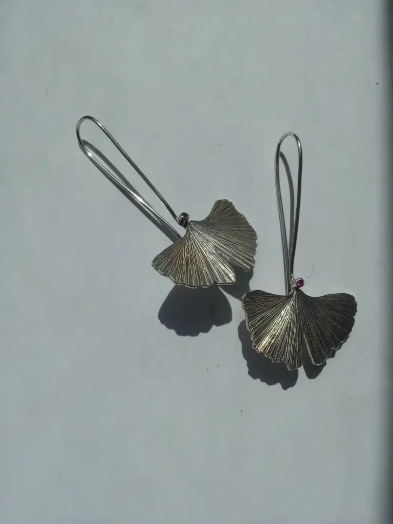 Silver earrings shaped like ginkgo leaves with a semi-circular clasp and small pink gemstones.