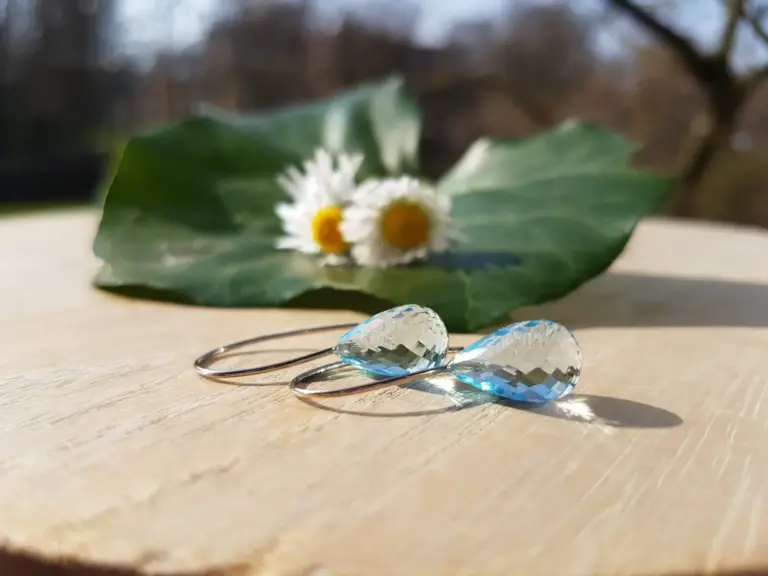 Two silver hoop earrings with large, teardrop-shaped, faceted clear blue stones resting on a light wooden surface. A green holly leaf with white flowers and yellow centers is in the background.