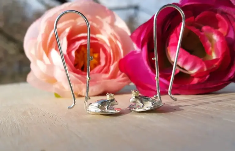 Silver earrings with small mouse figures on a wooden surface, with pink and red roses in the background.