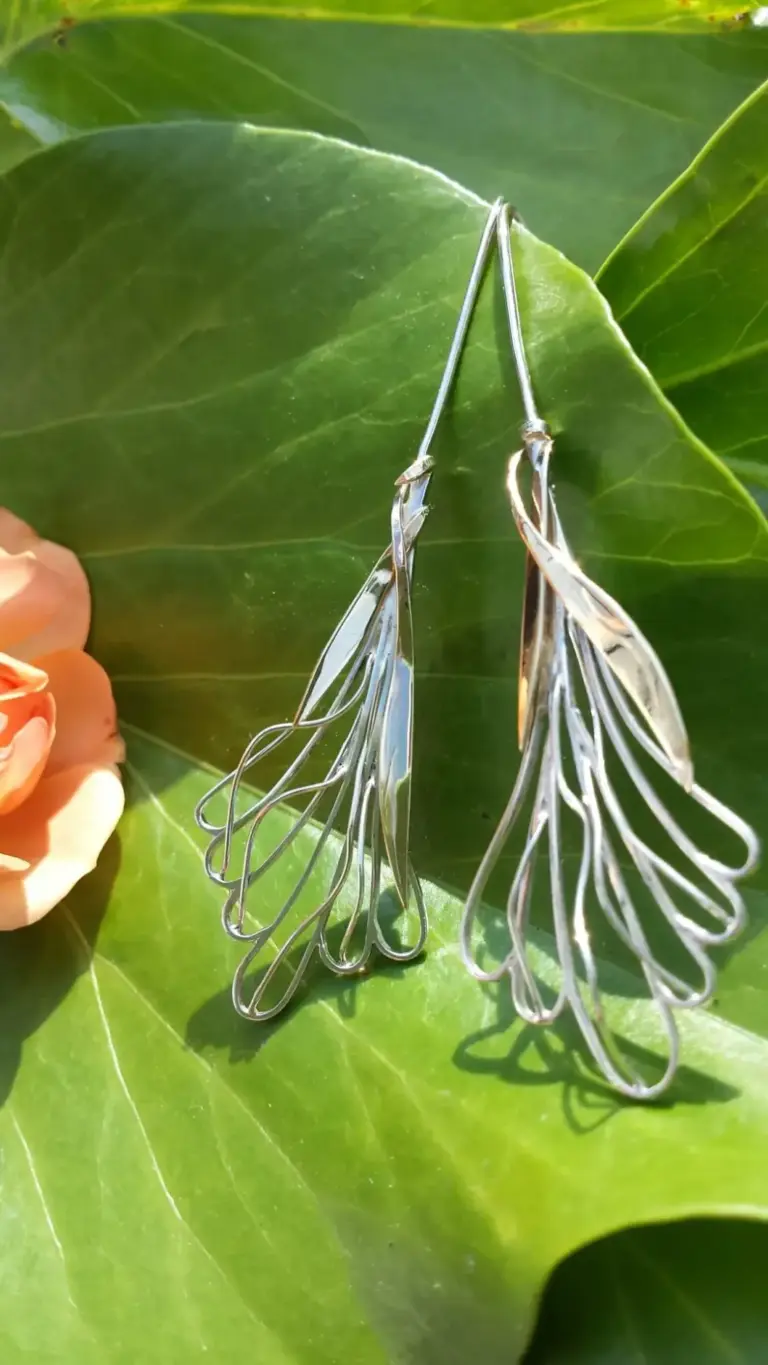 Close-up of silver jewelry with leaf-shaped pendants on large green leaves with a peach-colored flower in the corner.