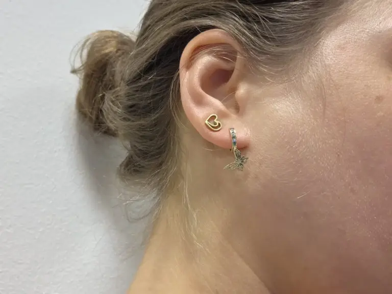 Close-up of a woman's ear with two earrings: a gold heart-shaped earring and a hoop earring with a butterfly charm.