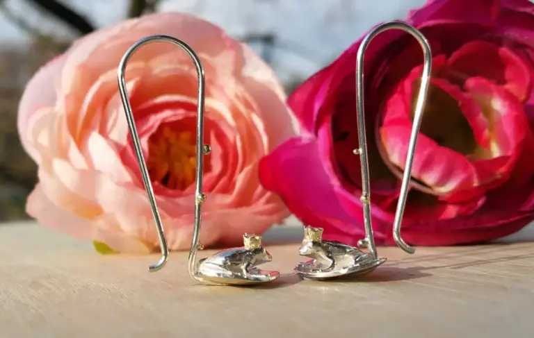 Close-up of two silver decorative paperclip holders shaped like birds, placed in front of large pink and red roses on a wooden surface.