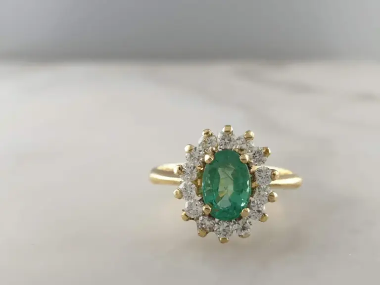 Close-up of a gold ring with a large green oval gemstone surrounded by smaller clear diamonds, set against a neutral background.