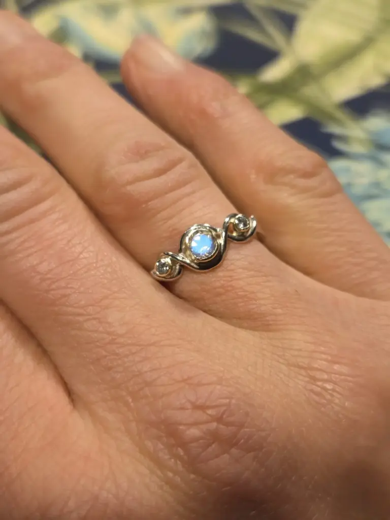 Close-up of a hand with a silver ring on the finger, featuring a light blue gemstone in the center and two small clear stones on each side.