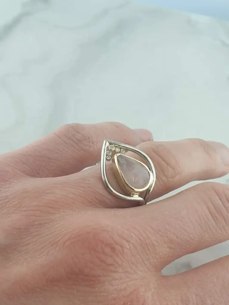 Close-up of a person's hand wearing a gold ring with a teardrop-shaped clear gemstone and small diamonds on the band.