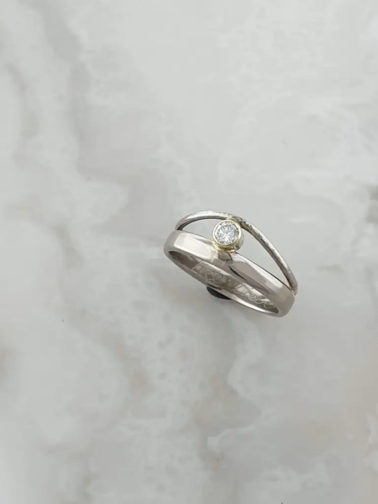 A silver ring with a single round diamond set in a yellow gold bezel on a white background.