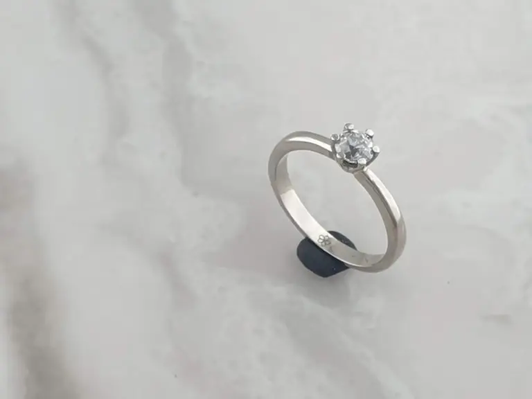 A silver engagement ring with a single round diamond in a prong setting, resting on a white surface.