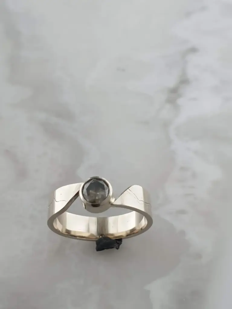 Metal ring with a dark gemstone on a silver base, placed on a white surface with a shadow nearby.