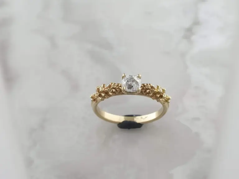 A gold ring with an intricate design and a large, clear gemstone in the center, displayed against a white background.