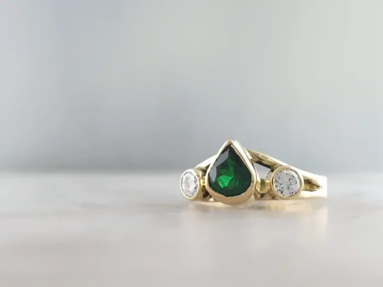 A gold ring with a large green gemstone surrounded by smaller clear gemstones, set against a plain light gray background.