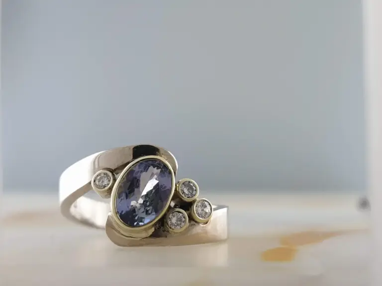 A silver ring with a large central purple gemstone surrounded by smaller clear gemstones, photographed against a gray background.