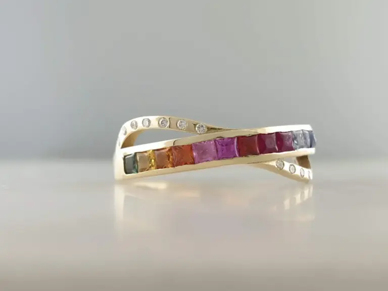 A gold ring with two rows of small clear gemstones, one row of round gemstones and one row of square rainbow-colored gemstones. The ring is positioned horizontally against a plain, light gray background.