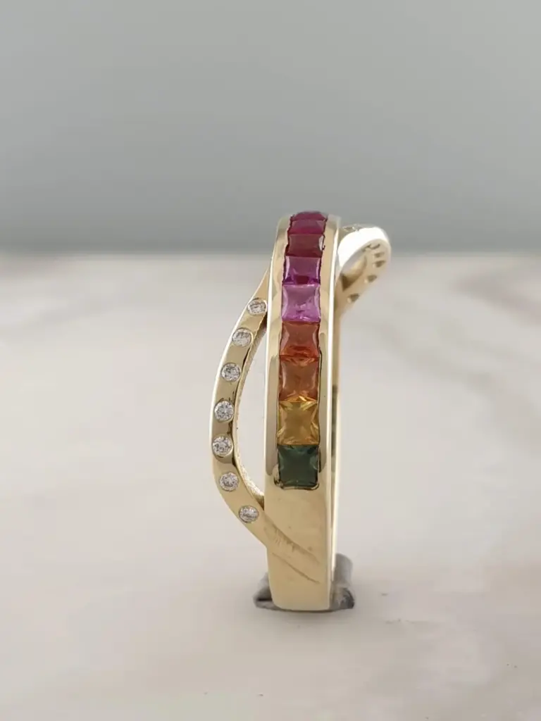 Two gold rings, each featuring a row of rectangular colored gemstones in red, pink, orange, yellow, green, and blue, are positioned against a plain background.