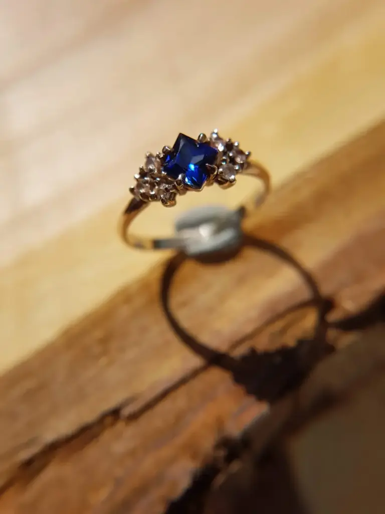 A close-up photo of a gold ring with a rectangular deep blue gemstone surrounded by small clear stones, resting on brown paper with handwritten text.