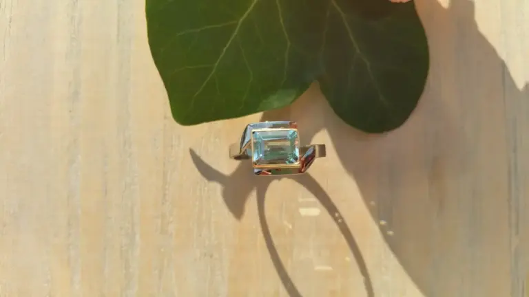 A silver ring with a large, rectangular blue gemstone set in the center, casting a shadow on a light-colored wooden surface. A large green leaf is partially visible in the background.