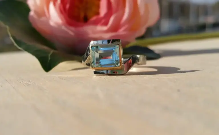 Close-up of a ring with a large square-cut blue gemstone, placed on a light surface with a pink flower in the background.