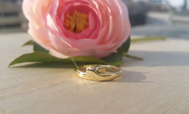 A gold heart-shaped ring placed on a light wooden surface in front of a pink and yellow flower with green leaves, with a blurred background.