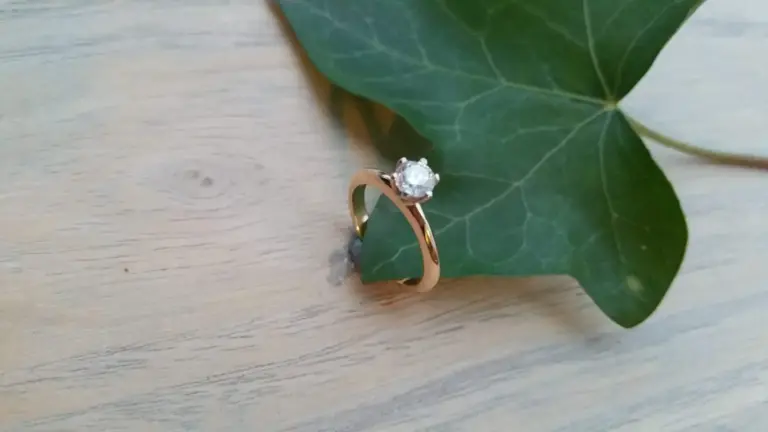 A gold ring with a large round diamond resting on a green ivy leaf on a light wooden surface.