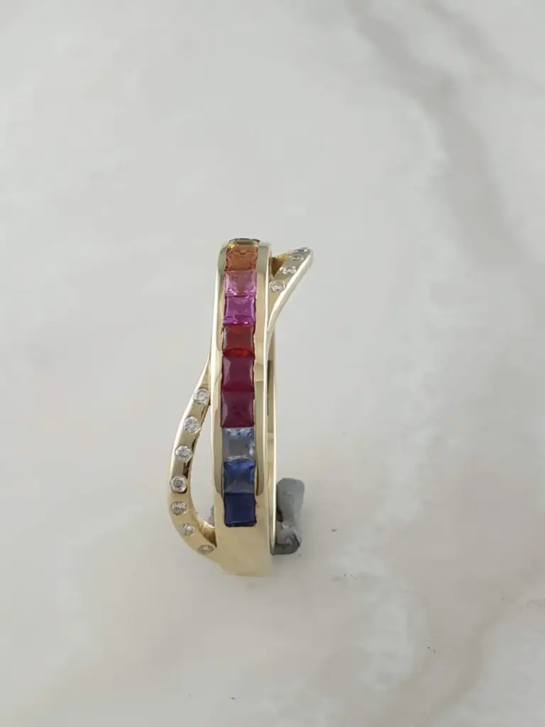 Close-up of colorful rings with gemstones, featuring rainbow-colored gemstones in a gold band against a plain light background.