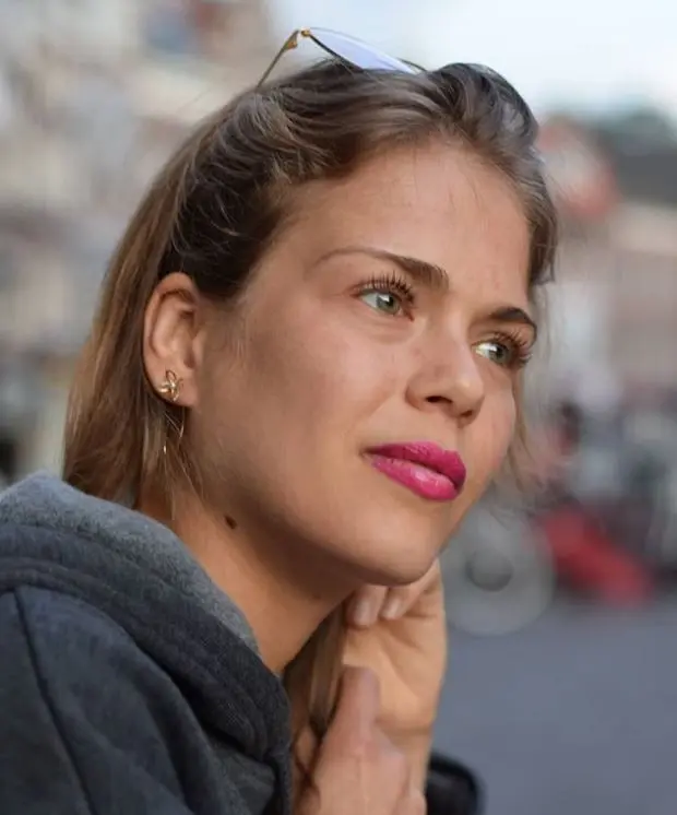 Young woman with brown hair, pink lipstick, and sunglasses on her head, wearing a gray hoodie, outdoors, with a blurred background.