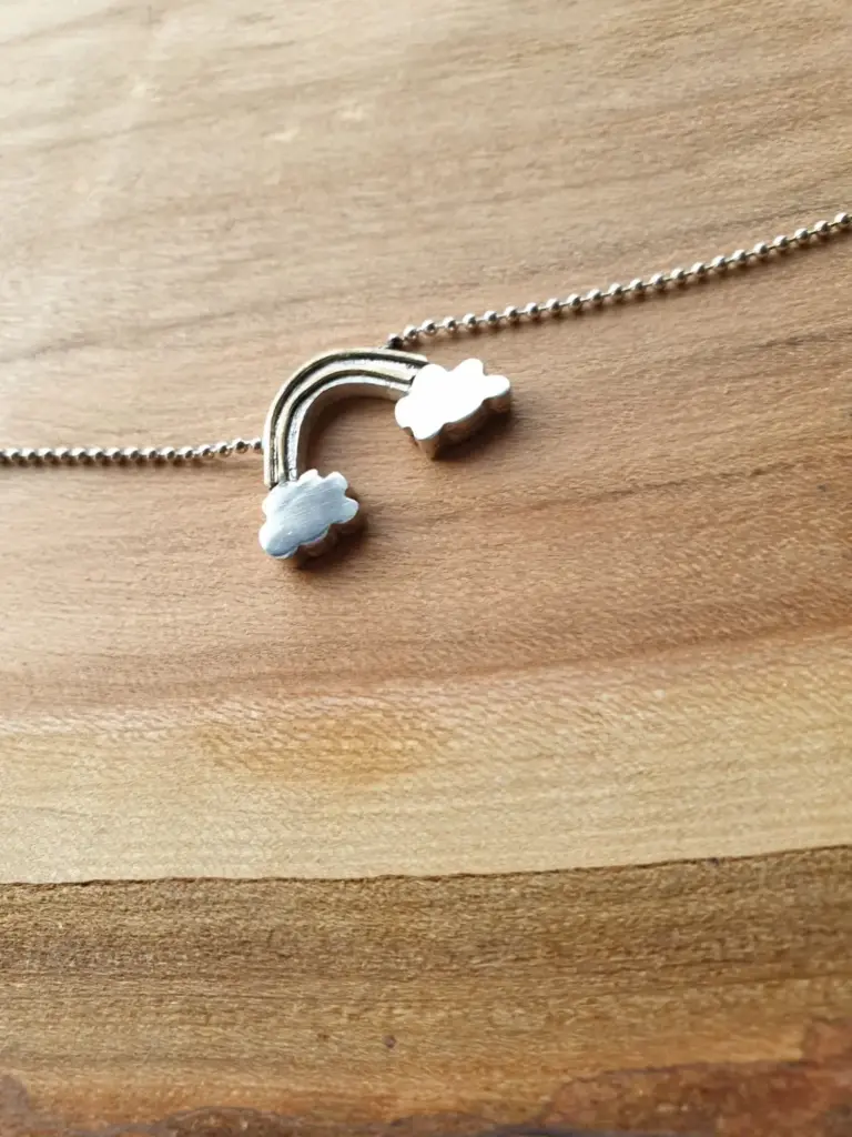 Close-up of a silver necklace with a rainbow and cloud pendant on a wooden surface.