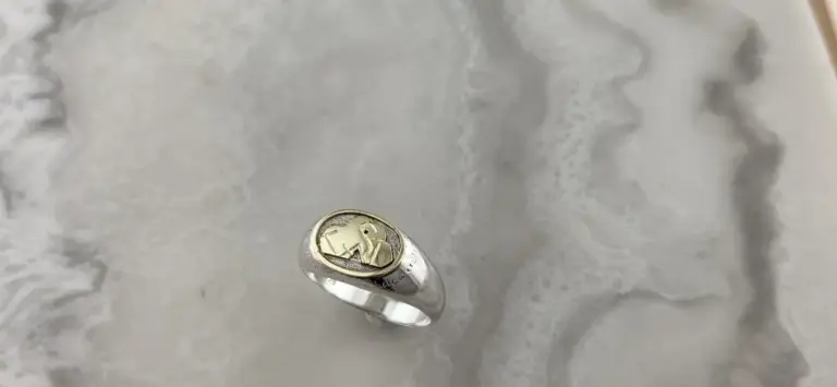 A silver ring with a gold and silver logo on top, featuring an eagle emblem, resting on a textured surface.